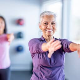 A smiling senior woman in a purple top leads an arm-stretching fitness class in a bright, modern studio.