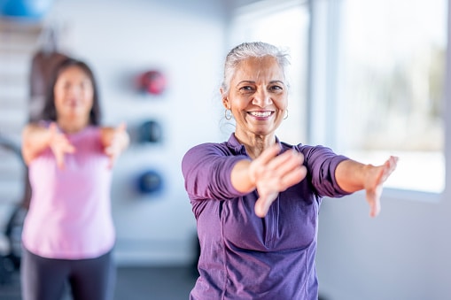 A smiling senior woman in a purple top leads an arm-stretching fitness class in a bright, modern studio.