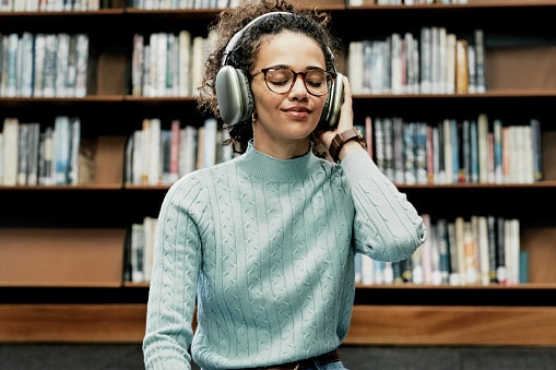 Happy woman, headphones and listening with audio book in library.